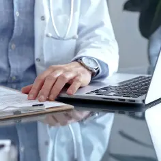 Lekarz w białym kitlu pisze na dokumentach, obok laptop. Może to być wizyta u psychiatry.