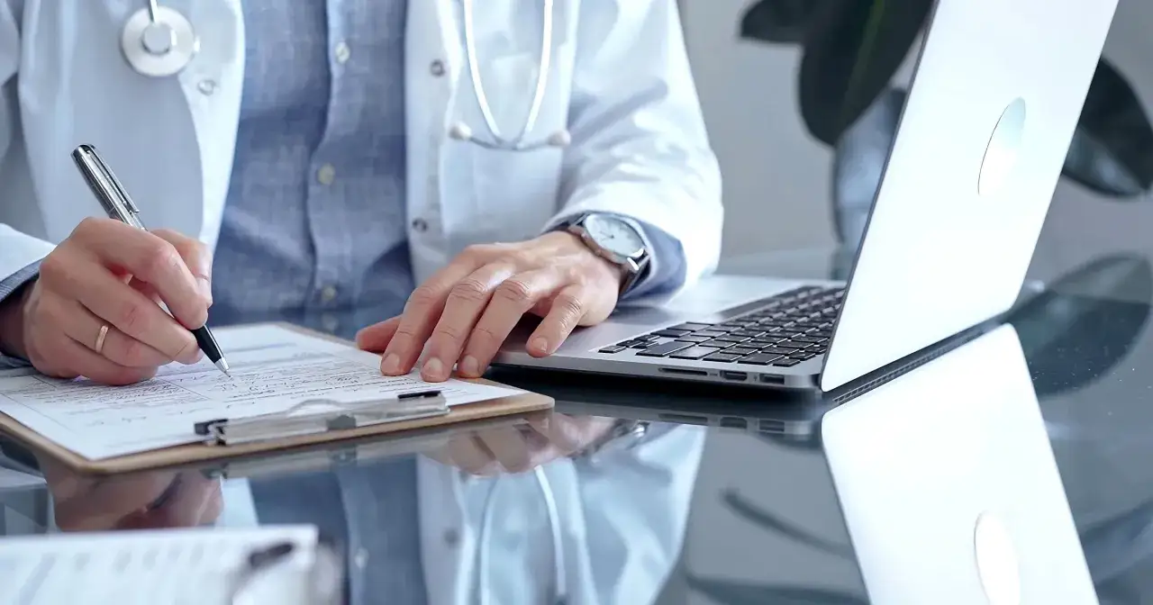 Lekarz w białym kitlu pisze na dokumentach, obok laptop. Może to być wizyta u psychiatry.